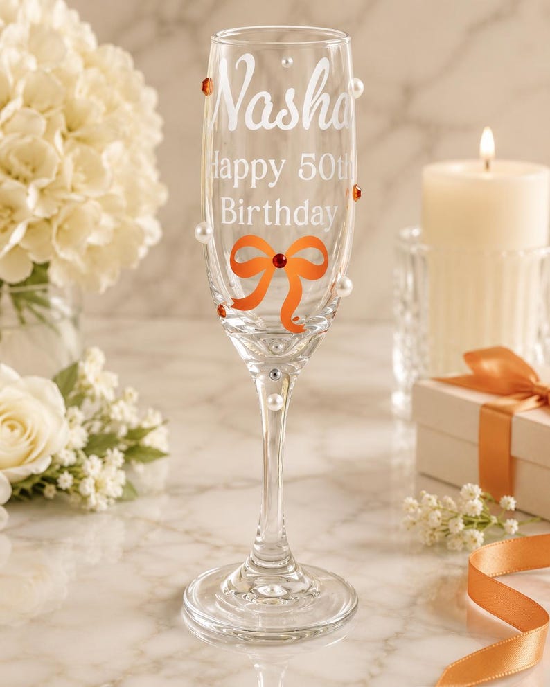 May include: A clear glass champagne flute featuring the white etched text Nasha Happy 50th Birthday. The glass is decorated with an orange bow graphic, small white pearls, and orange and silver rhinestones applied to the exterior surface.