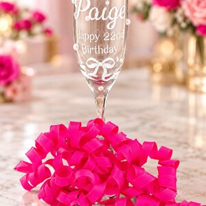 May include: A clear glass champagne flute featuring the white printed text Paige, Happy 22nd Birthday, and a white bow graphic. The glass is decorated with small white pearl accents and is surrounded by a bright pink curled ribbon at the base.
