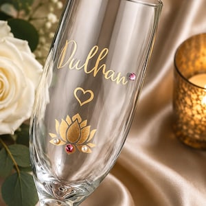 May include: A clear champagne flute with gold lettering that reads "Dulhan", a heart, and a gold lotus flower design. The lotus flower has a pink and two white gems. The glass is on a satin surface.