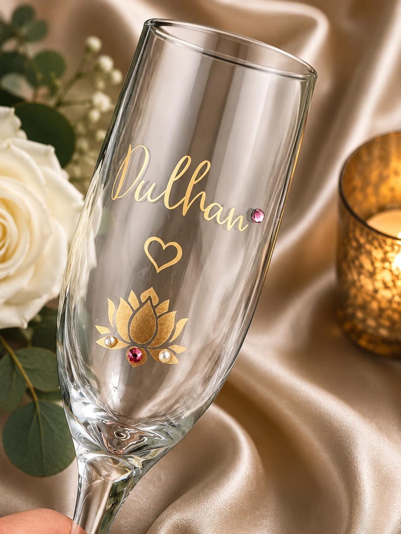May include: A clear champagne flute with gold lettering that reads "Dulhan", a heart, and a gold lotus flower design. The lotus flower has a pink and two white gems. The glass is on a satin surface.