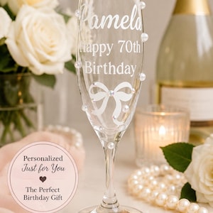 May include: A clear glass champagne flute featuring the white printed text Pamela, Happy 70th Birthday, and a white bow graphic. The glass is decorated with several small, round, iridescent white pearls attached to the exterior surface.