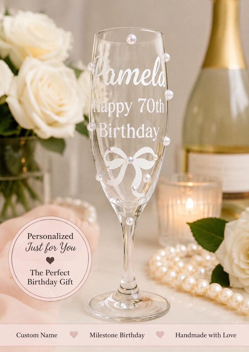 May include: A clear glass champagne flute featuring the white printed text Pamela, Happy 70th Birthday, and a white bow graphic. The glass is decorated with several small, round, iridescent white pearls attached to the exterior surface.