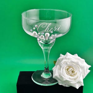 May include: A clear glass coupe cocktail glass featuring the name Cheri etched in script font on the side. The glass is decorated with various sizes of white pearls and iridescent rhinestones. A large white artificial rose sits beside the base.