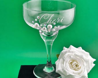 May include: A clear glass coupe cocktail glass featuring the name Cheri etched in script font on the side. The glass is decorated with various sizes of white pearls and iridescent rhinestones. A large white artificial rose sits beside the base.