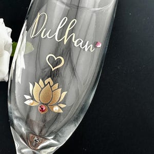 May include: A clear champagne flute with the name "Dulhan" in gold script. A gold lotus flower with a red gem and pearls is at the base. A heart and a pink gem are also on the glass.