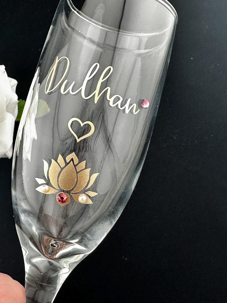 May include: A clear champagne flute with the name "Dulhan" in gold script. A gold lotus flower with a red gem and pearls is at the base. A heart and a pink gem are also on the glass.