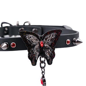 May include: Black choker necklace featuring a decorative butterfly pendant with red accents. The butterfly is embellished with a red gem in the center and a dangling teardrop gem. The choker has silver spikes and red gem studs.