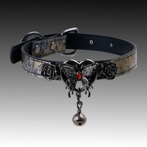 Könnte beinhalten: Ein schwarzes Halsband mit texturiertem Gold- und Schwarzmuster, verziert mit zwei schwarzen Rosen und einem zentralen schwarzen Schmetterlingsanhänger mit rotem Stein. Eine kleine, runde Glocke aus dunklem Metall hängt am Schmetterling.