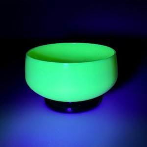 May include: A luminous, neon green glass bowl with a rounded form. The bowl rests on a dark, circular base. The background is a gradient from dark blue to black.