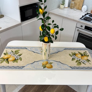 Op de afbeelding: Een witte tafel met een beige tafelloper met citroen- en bloemmotieven. Een vaas met een citroentak staat in het midden van de tafel. De keukenomgeving omvat witte kasten en apparaten.