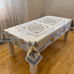 Könnte beinhalten: Rechteckige Tischdecke mit beigem Hintergrund und einem sich wiederholenden Muster aus Zitronen und Blättern. Der Rand hat ein blau-weißes Design. Die Mitte der Tischdecke hat zwei große quadratische Medaillons mit einem blau-gelben Blumendesign.