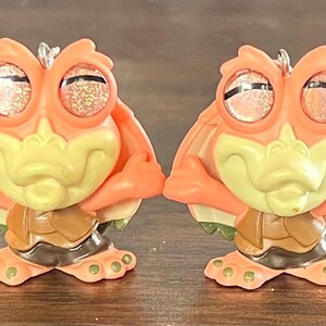 May include: Two orange and yellow cartoon frog figurines with glittery eyes and a brown shell-like base. The frogs have their arms folded and are standing upright. The background is a dark brown surface.