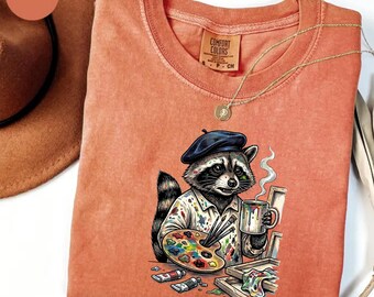 May include: Orange Comfort Colors t-shirt featuring a raccoon artist wearing a blue beret and holding a coffee mug. The raccoon is holding a paint palette and paintbrushes. The shirt has a round neck and a Comfort Colors tag.
