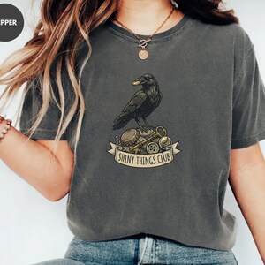 Puede incluir: Camiseta gris oscuro con un cuervo negro sosteniendo una moneda de oro, posado sobre llaves, tapones de botella y botones. La pancarta de abajo dice "Shiny Things Club". La camiseta es de estilo casual.