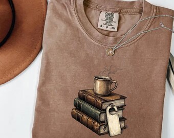 Puede incluir: Una camiseta marrón Comfort Colors con un gráfico de una taza de café sobre una pila de libros. Un collar plateado con un colgante está en la camisa. La camisa tiene una pequeña etiqueta que dice S-CH.