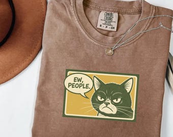 May include: Espresso-colored Comfort Colors t-shirt with a graphic of a grumpy cat and the words "Ew, People" in a speech bubble. The shirt has a crew neck and short sleeves. A silver necklace with a small pendant is on the shirt.