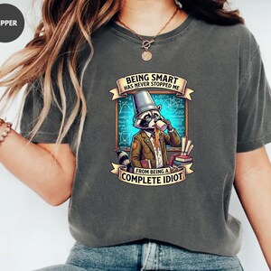 Puede incluir: Camiseta gris con un mapache de dibujos animados bebiendo café, con el texto "BEING SMART HAS NEVER STOPPED ME FROM BEING A COMPLETE IDIOT." El mapache lleva un sombrero y una chaqueta, con un fondo de pizarra.