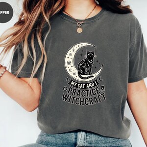 May include: A charcoal grey short-sleeved t-shirt featuring a graphic print of a black cat sitting inside a crescent moon. The design includes small star and pentagram symbols, with the text "MY CAT AND I PRACTICE WITCHCRAFT" printed below.