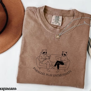 Könnte beinhalten: Espressofarbenes T-Shirt mit einer Grafik von zwei Waschbären auf einem Sofa, einer liest und der andere benutzt einen Laptop. Der Text "PARALLEL PLAY ENTHUSIASTS" steht darunter. Eine silberne Halskette und ein brauner Hut sind ebenfalls zu sehen.