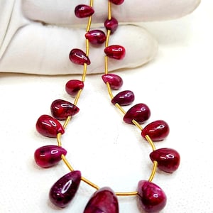 May include: A necklace with teardrop-shaped, deep red gemstones strung on gold-coloured wire. The gemstones have a glossy finish and vary slightly in size. The background is a white surface.