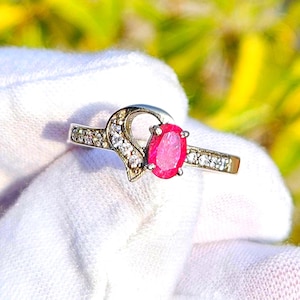 May include: A silver ring featuring a vibrant oval-cut ruby gemstone as the focal point. The ring is adorned with smaller, clear gemstones along the band and a decorative heart-shaped design, creating a sparkling and elegant appearance.