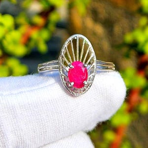 Puede incluir: Un anillo de plata con una gema ovalada de color rosa brillante. La gema está engastada en un marco calado. El anillo se muestra sobre un fondo blanco, con un fondo borroso verde y amarillo.
