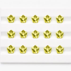 May include: Fifteen faceted star-shaped gemstones in a bright lemon yellow color. The stones are arranged in three horizontal rows of five on a white display board with horizontal ridges, showcasing a clear, sparkling, and geometric cut.