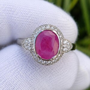 May include: A silver ring featuring a large, oval-cut ruby gemstone. The ruby is surrounded by a halo of small, clear diamonds, with additional diamond clusters on each side of the main stone. The ring is set against a white background.