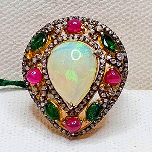 May include: A gold ring with a pear-shaped opal centre stone, surrounded by small diamonds, red rubies, and emerald green gemstones. The ring's design is intricate and detailed, showcasing a luxurious aesthetic. The ring's design is intricate and detailed.