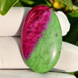 Puede incluir: Gema ovalada con una división de color vibrante. Una mitad es de un rojo intenso, mientras que la otra es de un verde brillante. La superficie de la piedra tiene una textura pulida y suave, con patrones naturales visibles.