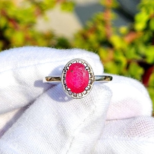 May include: A silver ring featuring a vibrant, oval-cut ruby gemstone. The ruby is a deep pink-red color, set within a decorative, textured bezel. The ring's band is slender and polished, creating a classic and elegant design.