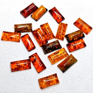 May include: A collection of rectangular amber cabochons featuring various shades of orange, brown, and deep gold. Each piece displays natural internal inclusions and a smooth, polished surface with a glossy finish, arranged in a scattered pattern.