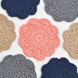 May include: A collection of handmade crochet coasters in various colors, including navy blue, gray, peach, and beige. Each coaster has a scalloped edge and a circular design, perfect for protecting surfaces from drink condensation.