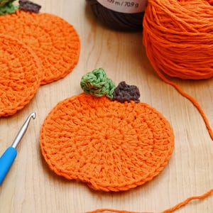 May include: Handmade orange crochet pumpkins with green and brown detailing, ideal for autumn decorations. The image also shows a blue crochet hook and orange yarn, suggesting a craft project. The pumpkins are round and have a textured appearance.