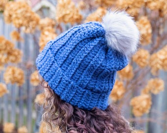 May include: A blue knitted beanie with a ribbed design and a fluffy, white pom-pom on top. The hat is made of a soft, textured material. The background features blurred, dried flowers.