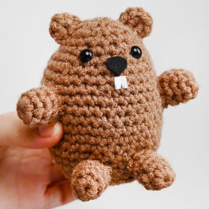 May include: A brown crochet beaver toy with black eyes, a black nose, and two white teeth. The beaver has small arms and legs. The toy is held against a white background.