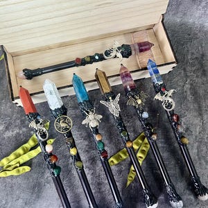 May include: A collection of handcrafted wands with crystal tips and decorative embellishments. Each wand features a unique crystal color, including red, white, blue, and purple, and is adorned with metal charms and colorful beads. The wands are displayed in a wooden box.
