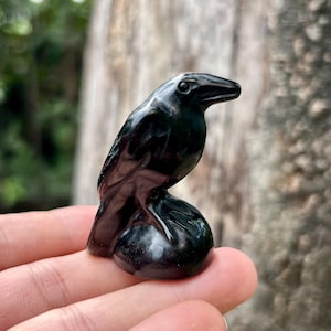 Puede incluir: Una figura de cuervo verde oscuro y brillante, posada sobre una base redondeada. El ave está tallada con plumas detalladas y un pico afilado. La escultura se sostiene en una mano, con un fondo natural borroso.