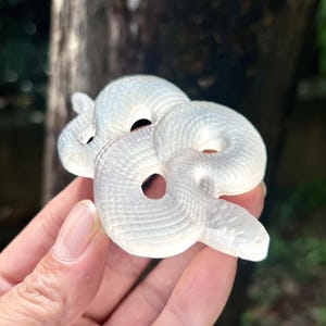 Puede incluir: Una figura de serpiente blanca enroscada con patrones de escamas detallados. La escultura tiene dos agujeros para los ojos y un agujero que atraviesa el cuerpo. La cabeza de la serpiente está ligeramente levantada.