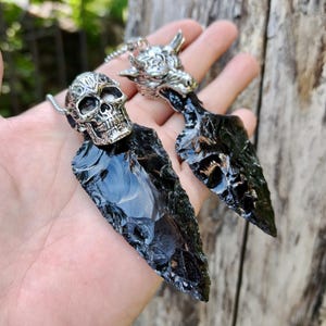 May include: Two pendants featuring black obsidian arrowheads. One has a silver skull, the other a silver wolf head. The obsidian is polished and reflective. The pendants are on a silver chain.