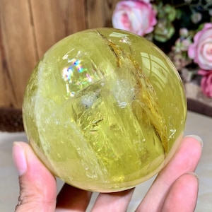 May include: A translucent, yellow-green crystal sphere. The sphere has internal fractures and inclusions, with iridescent flashes visible on its surface. The background includes wooden elements and pink roses, suggesting a decorative or collectible item.