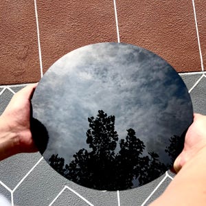 May include: A circular, black, reflective disc with a smooth, glossy surface. The disc acts as a mirror, displaying a clear reflection of the sky with clouds and the silhouettes of tree branches across its dark, polished, and circular face.