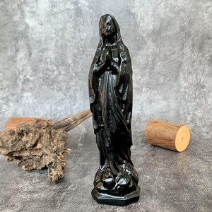 May include: A black statue of a religious figure, with hands clasped in prayer, stands on a decorative base. The figure is draped in flowing robes. The statue is set against a gray background with natural wood elements.