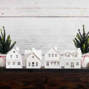 Puede incluir: Cinco casas de papel blanco con una cinta roja y blanca a rayas alrededor de la base de cada casa. Las casas est&aacute;n dispuestas en fila sobre una superficie de madera marr&oacute;n oscuro. Hay dos peque&ntilde;os &aacute;rboles de Navidad verdes en macetas blancas a cada lado de las casas.