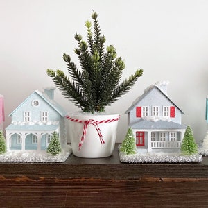 Puede incluir: Cinco casas en miniatura de papel mach&eacute; con techos cubiertos de nieve y vallas blancas, decoradas en colores pastel, est&aacute;n dispuestas en un estante de madera con peque&ntilde;os &aacute;rboles de hoja perenne en maceta.