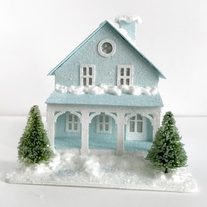 Puede incluir: Una casa de papel mach&eacute; azul claro con molduras blancas y un techo nevado. La casa tiene dos ventanas y una ventana redonda en la parte delantera. Hay dos peque&ntilde;os &aacute;rboles verdes a cada lado de la casa.