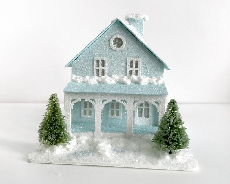 Puede incluir: Una casa de papel mach&eacute; azul claro con molduras blancas y un techo nevado. La casa tiene dos ventanas y una ventana redonda en la parte delantera. Hay dos peque&ntilde;os &aacute;rboles verdes a cada lado de la casa.