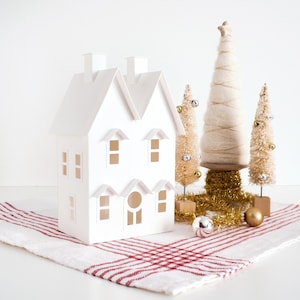 May include: A white paper house with cut-out windows and chimneys, set on a red and white striped cloth. Decorative Christmas trees and ornaments, including gold and silver baubles, complete the festive scene.