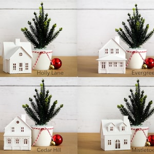 Puede incluir: Cuatro casas de papel blanco con una cuerda roja y blanca alrededor de la base de un peque&ntilde;o &aacute;rbol de Navidad verde en maceta. Cada casa tiene un nombre diferente: Holly Lane, Evergreen Way, Cedar Hill y Mistletoe Place.