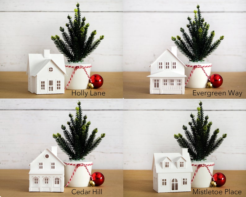 Puede incluir: Cuatro casas de papel blanco con una cuerda roja y blanca alrededor de la base de un peque&ntilde;o &aacute;rbol de Navidad verde en maceta. Cada casa tiene un nombre diferente: Holly Lane, Evergreen Way, Cedar Hill y Mistletoe Place.